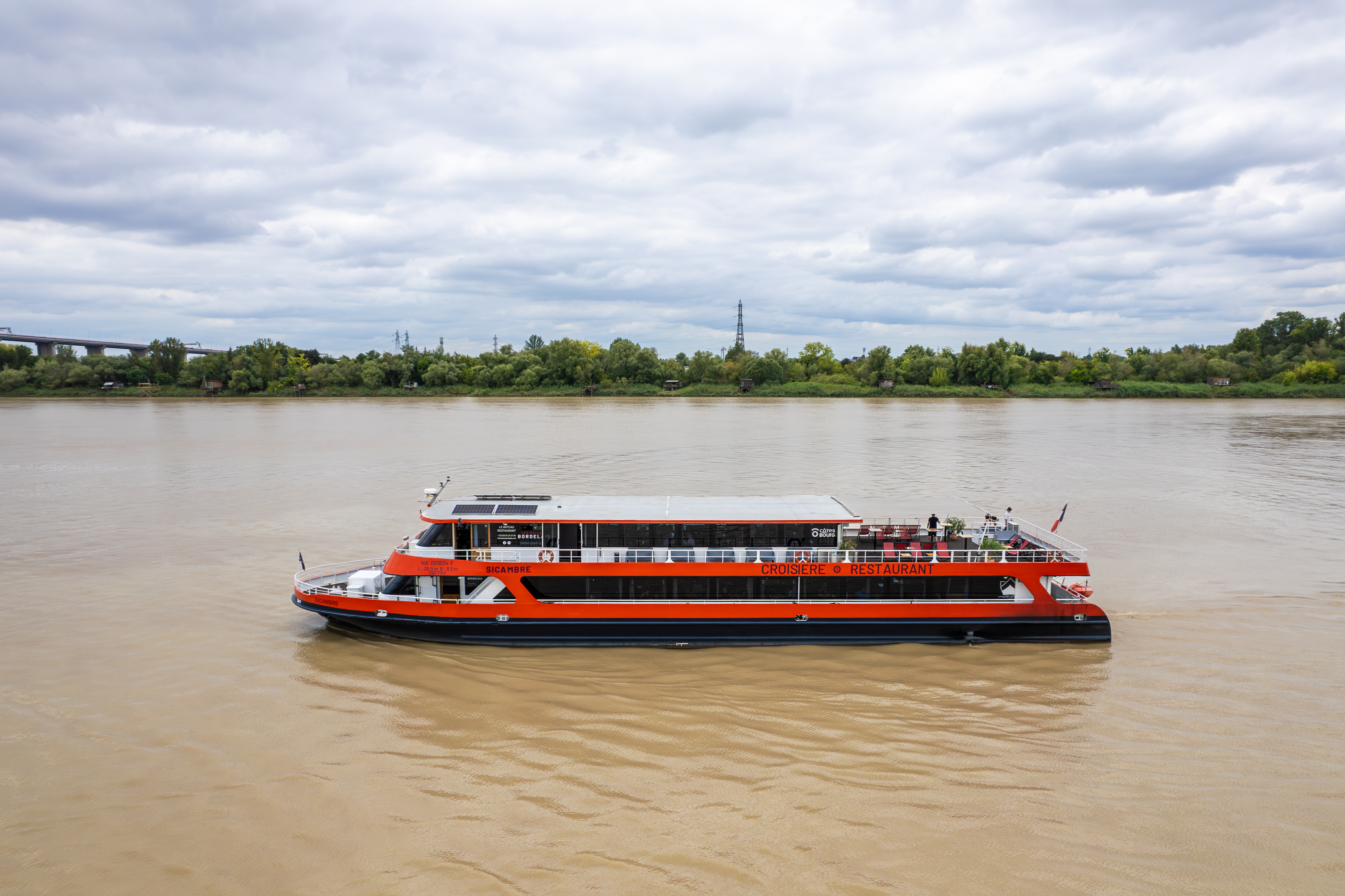 Ein rot-weißes Kreuzfahrtschiff fährt auf der Garonne in Bordeaux, mit grünen Ufern und bewölktem Himmel im Hintergrund.