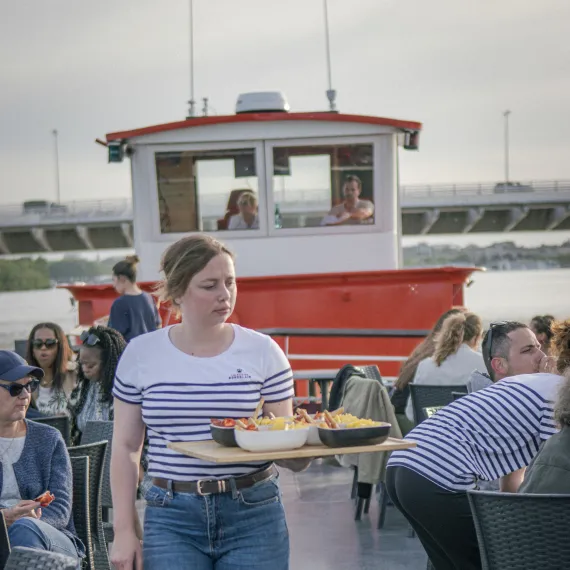 Serveuse apportant des tapas à bord d'une croisière sur la Garonne à Bordeaux, avec les passagers installés sur le pont du bateau.