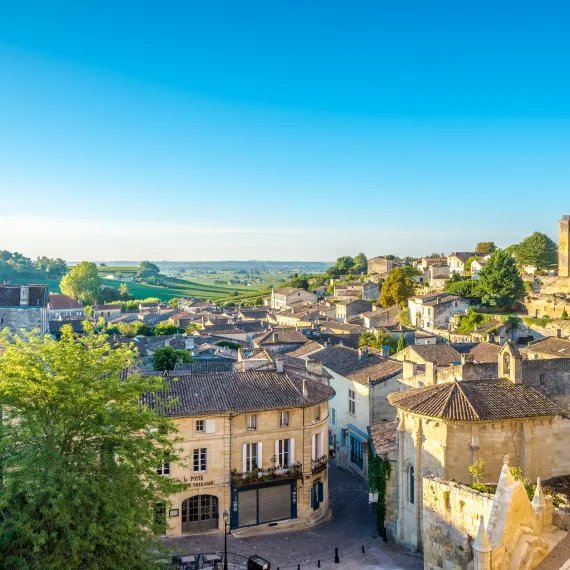 Vista panorâmica de Saint-Émilion com seus edifícios medievais em pedra, a característica igreja monolítica, telhados de telha vermelha e vinhedos da região de Bordéus estendendo-se até ao horizonte sob um céu azul claro.