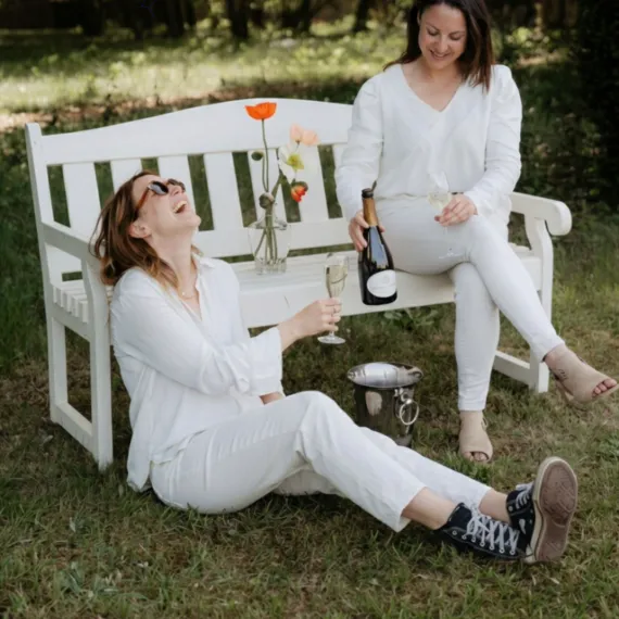 Deux femme dégustant une bouteille de vin effervescent au pied d'un banc dans un cadre verdoyant.