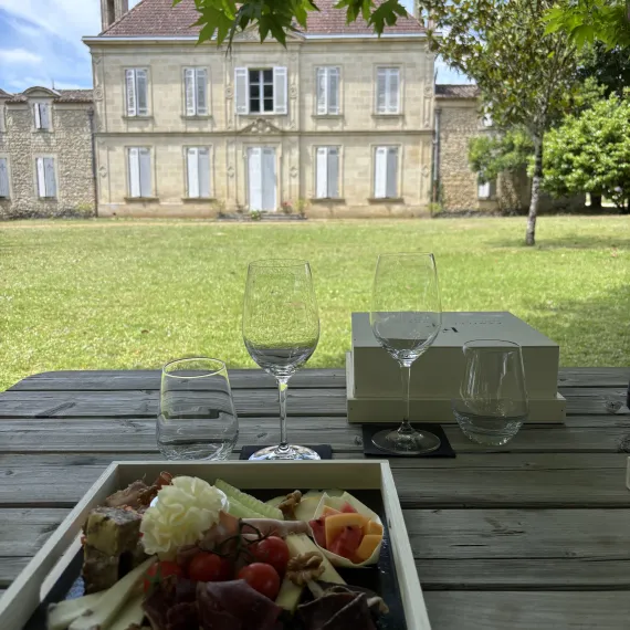 Verres à vin et planche apéritive dressés sur une table en bois face au Château Mangot, dans le parc arboré.