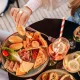 Main se servant dans un assortiment de mets sucrés et salés lors d'un brunch-croisière à Bordeaux, avec viennoiseries, fromages, charcuterie, tartines, fruits frais et boissons.