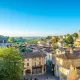 Vista panorâmica de Saint-Émilion com seus edifícios medievais em pedra, a característica igreja monolítica, telhados de telha vermelha e vinhedos da região de Bordéus estendendo-se até ao horizonte sob um céu azul claro.