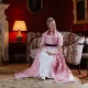 Femme en costume d'époque assise dans un salon historique aux murs rouges.